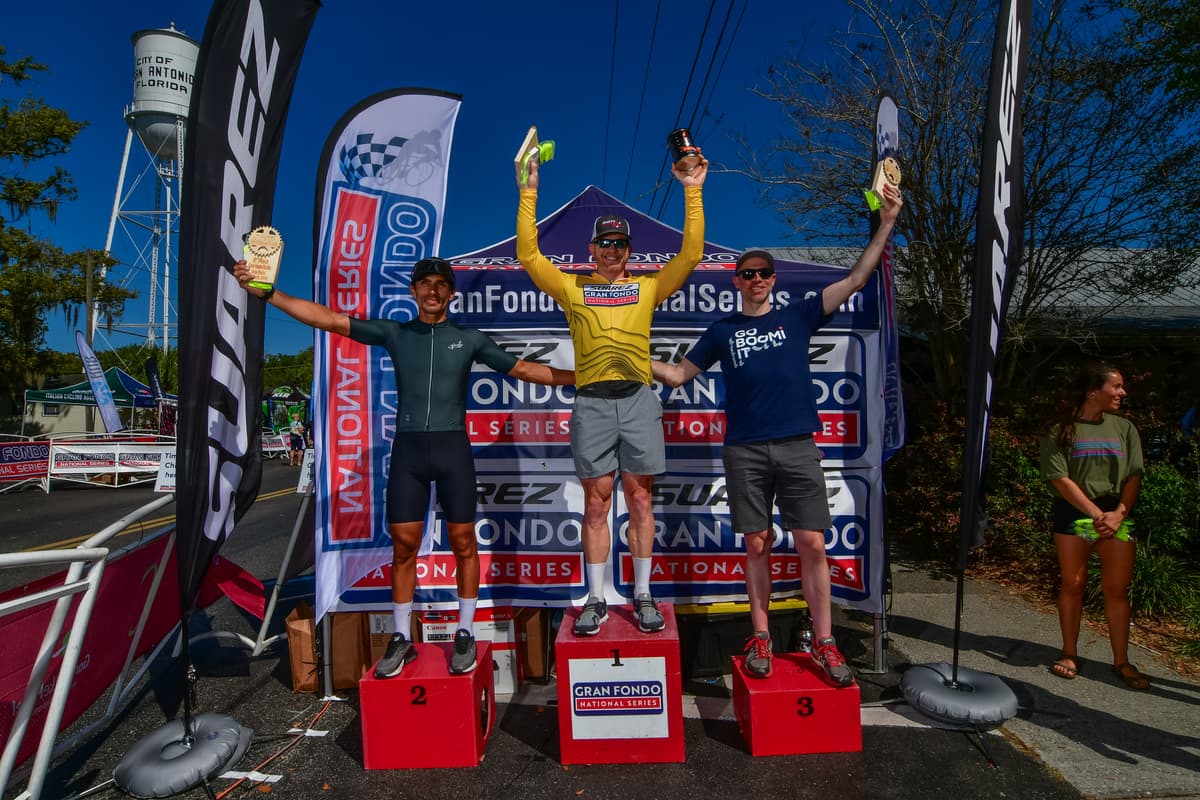 Three riders on the Gran Fondo National Series podium in San Antonio, Florida, arms raised with their trophies, the winner in the yellow leader's jersey on the top step.